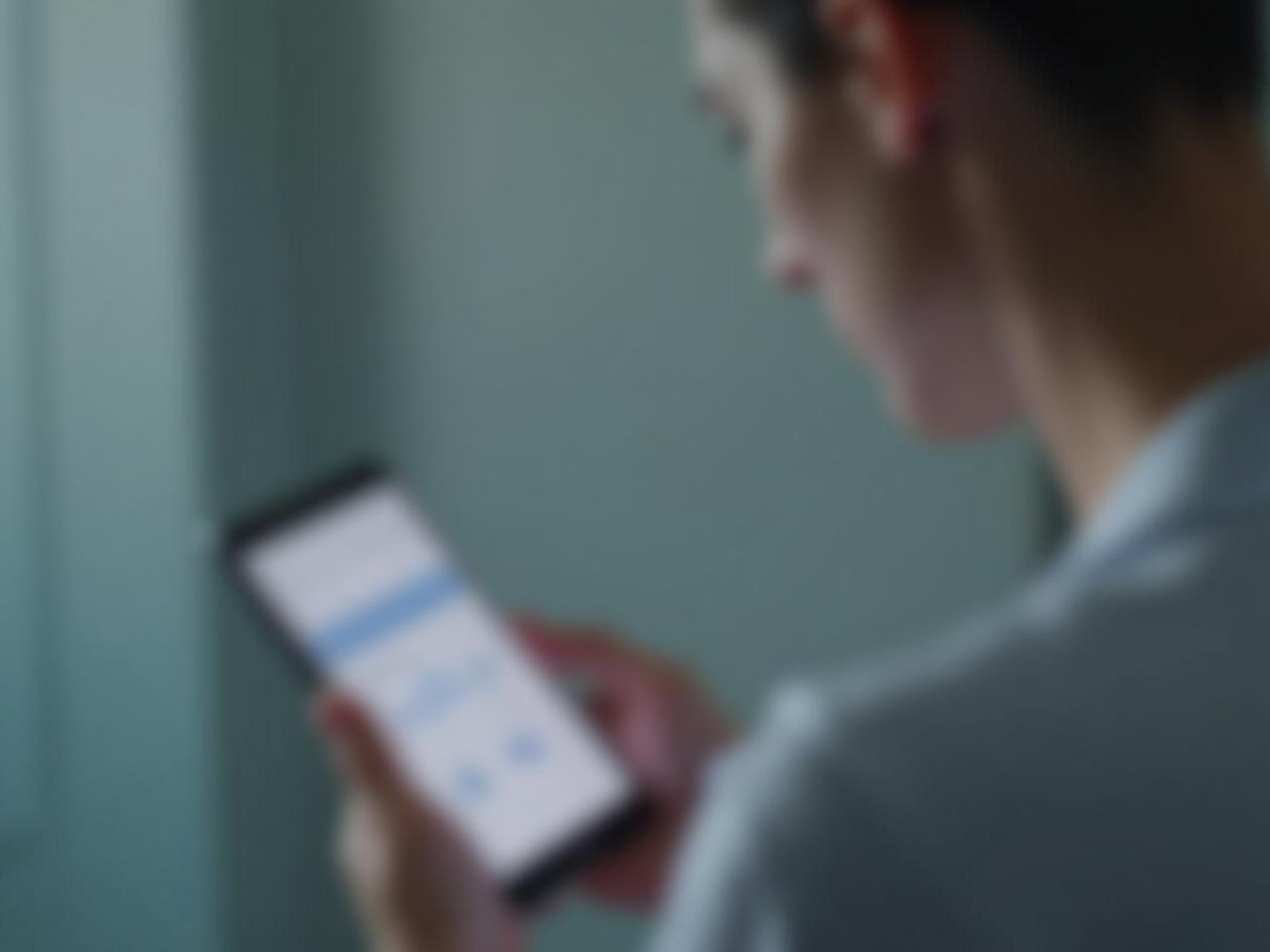 A close-up of a person looking thoughtfully at a smartphone displaying health data, with a background blurred to suggest a medical or technological environment.